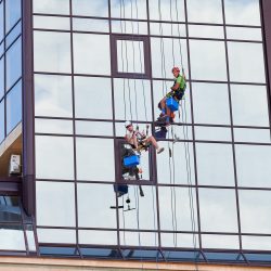 Industrial mountaineering workers washing glass windows of high-rise building, hanging on safety climbing ropes. Two men window cleaners working together outside skyscraper.